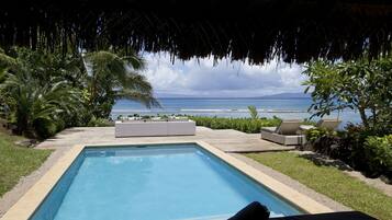 Une piscine avec vue sur l’océan, entourée de verdure et surmontée d’un toit de chaume.