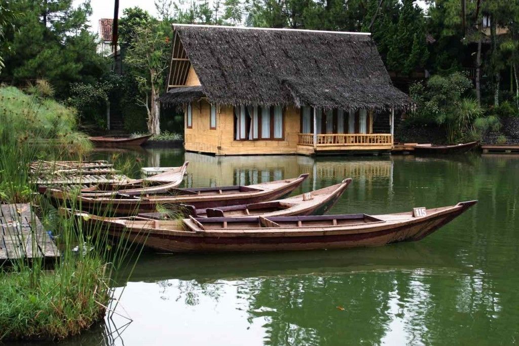 A thatched-roof house with a wooden deck, surrounded by calm water and several wooden boats.