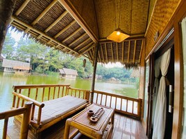 Una terraza de madera con techo de paja, con vistas a un río tranquilo y casas tradicionales al fondo.