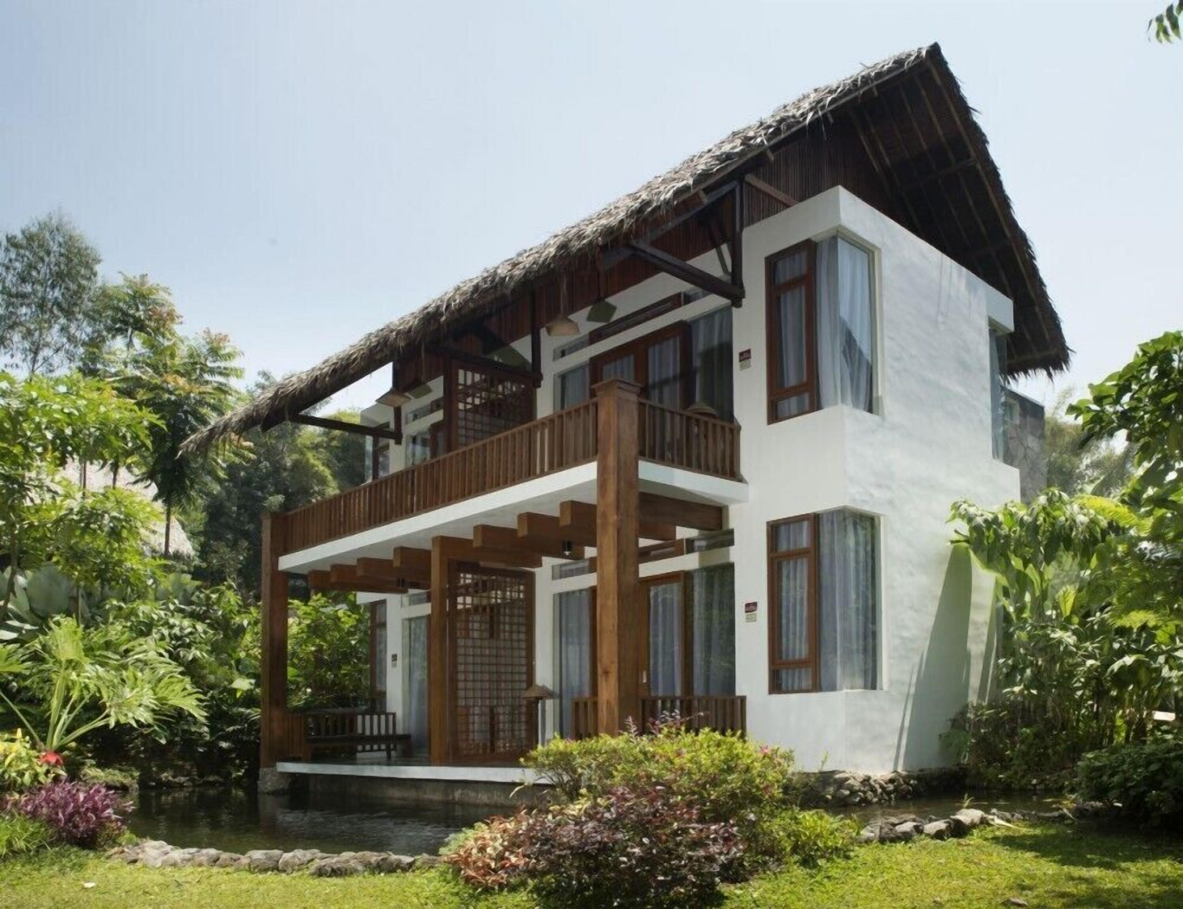 Una casa de dos pisos con techo de paja, balcón de madera y un jardín con un estanque.