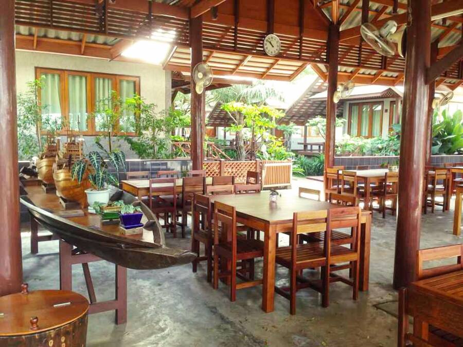 A covered outdoor dining area with wooden tables and chairs, surrounded by potted plants and a view of a traditional building.