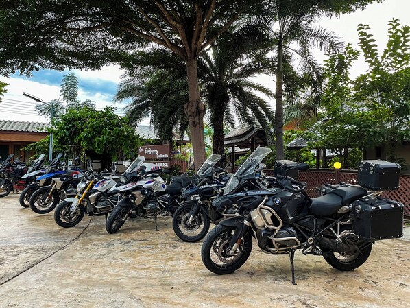 A row of motorcycles parked outdoors, with trees and a building in the background. - Baan Siriporn Resort (Samut Songkhram)