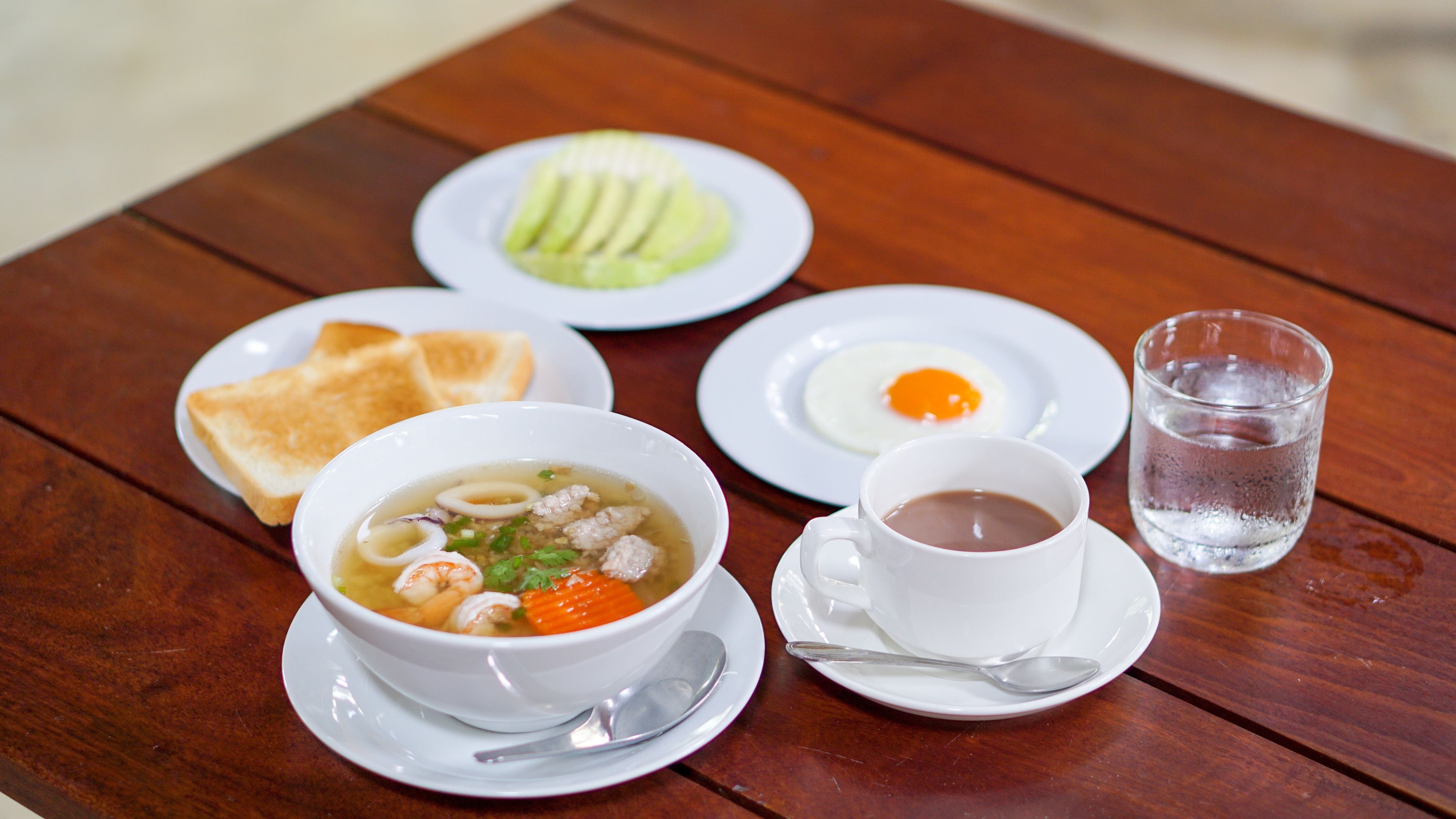 A table with a bowl of soup, a plate of sliced avocado, a plate with an egg, a glass of water, and a cup of tea.