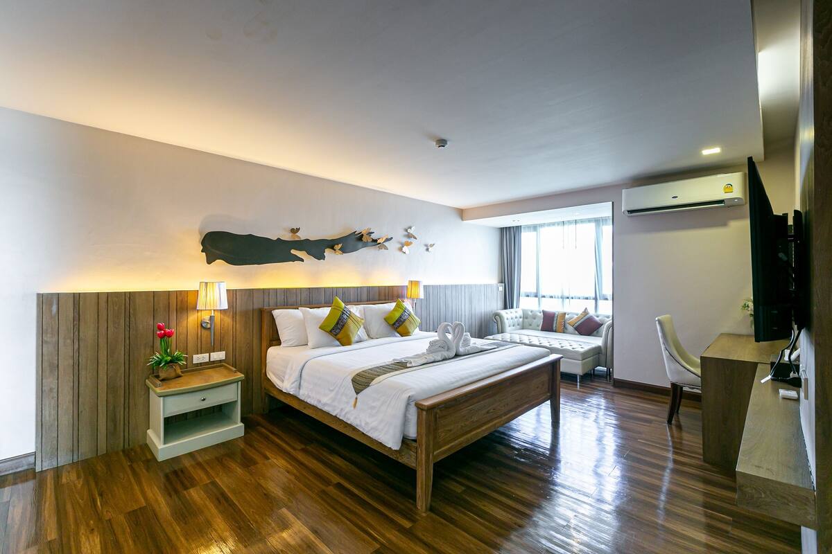 A modern hotel room with a wooden floor, a large bed with white bedding, a nightstand with a lamp, a desk, and a chair.