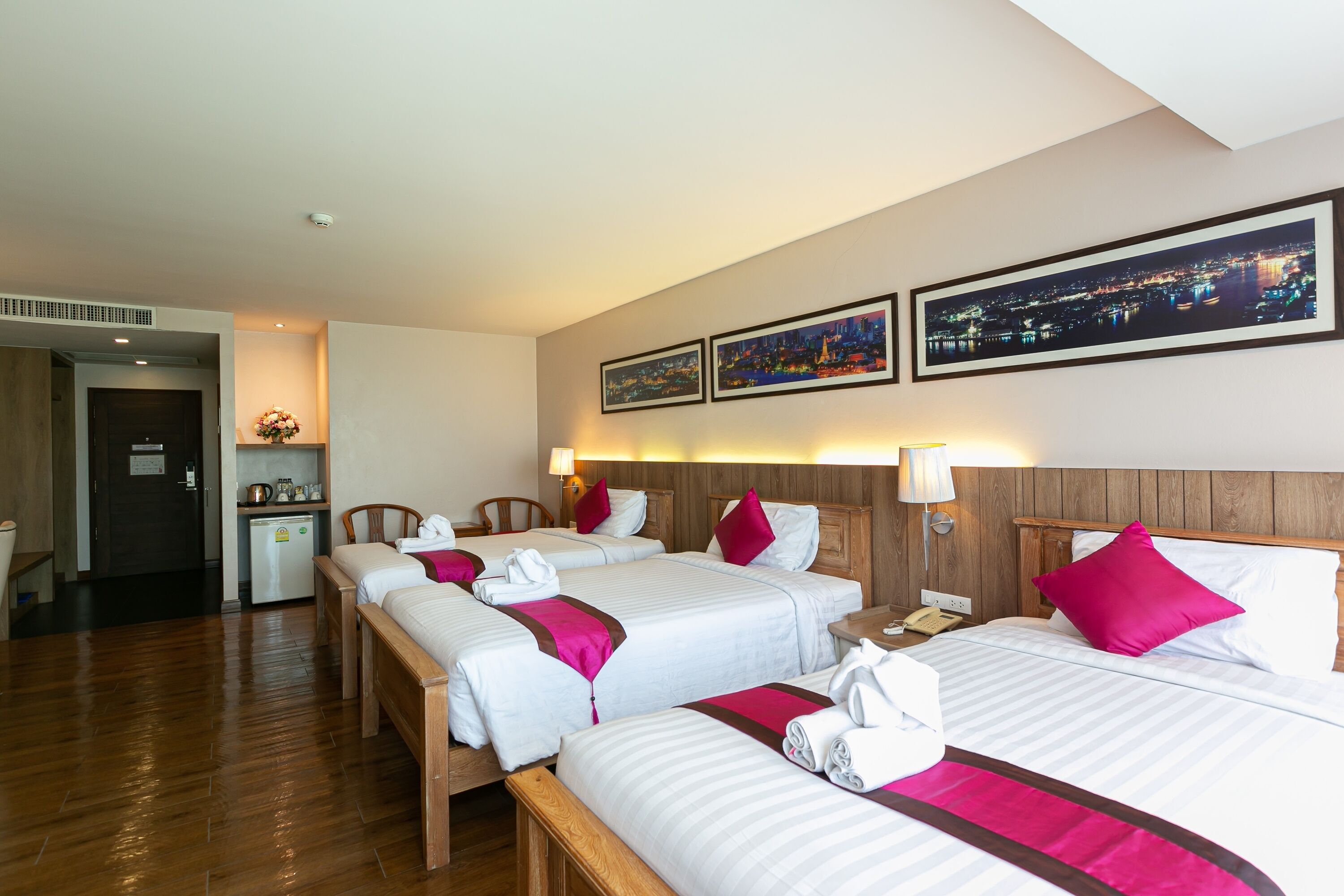 A hotel room with four beds, a wooden floor, a small kitchenette, and two framed cityscape pictures on the wall.