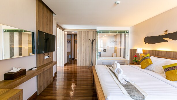 A hotel room with a wooden floor, a bed with white linens, a bedside table, a TV mounted on the wall, and a mirror. - Grand Ratchapruek Hotel (Pak Kret)