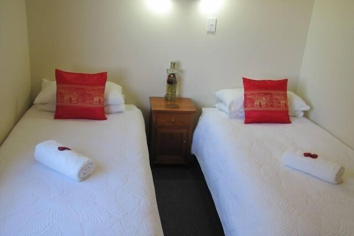 Ein Hotelzimmer mit zwei Betten, jedes mit einem roten Kopfkissen und einem zusammengerollten Handtuch. Dazwischen steht ein Nachttisch aus Holz mit einer Flasche darauf.
