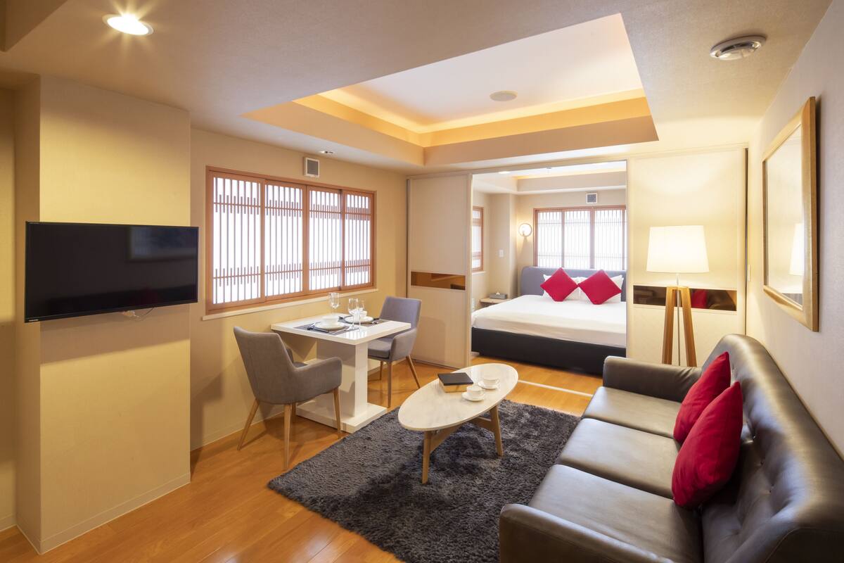A modern hotel room with a dining area, a sofa, a TV, and a bed with red pillows.