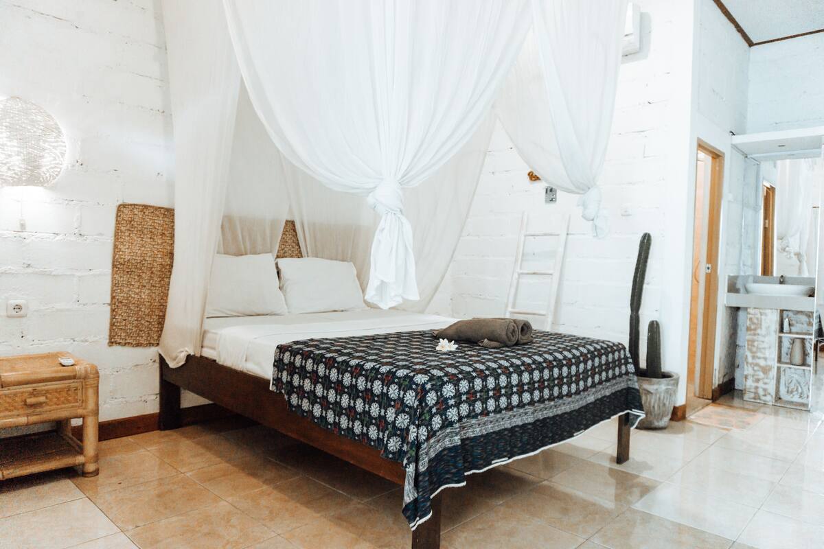 A bedroom with a bed, a nightstand, a woven wall hanging, a cactus, and a white door.