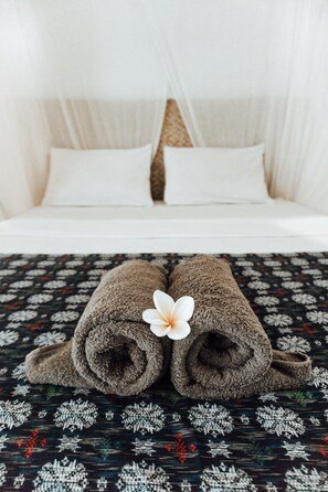 A bed with white linens and a canopy, a patterned bedspread, and two rolled towels with a white flower. - Balangan Inn (Jimbaran)