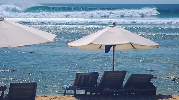 A beach with three lounge chairs and two umbrellas, facing the ocean with waves crashing in the background.