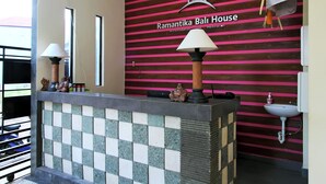 Un área de recepción con pared de baldosas a cuadros, un mostrador de recepción con una lámpara y un cartel en la pared que dice "Ramantika Bali House".