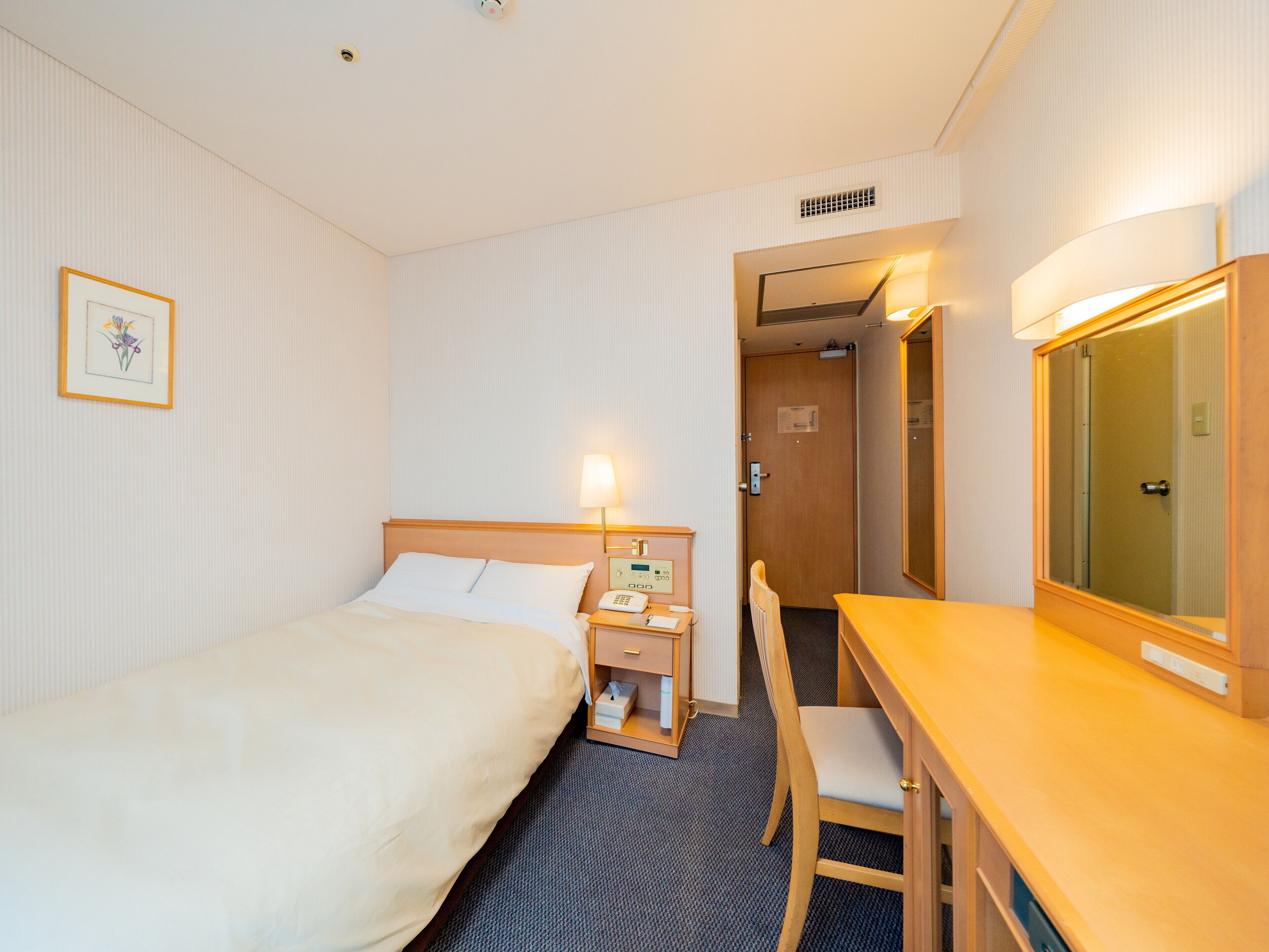 A hotel room with a bed, bedside table, chair, desk, lamp, and a mirror.