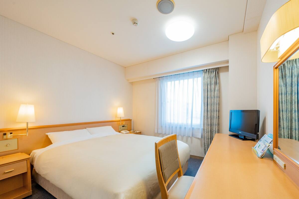 A hotel room with a large bed, bedside tables, a desk with a chair, a television, and a window with curtains.