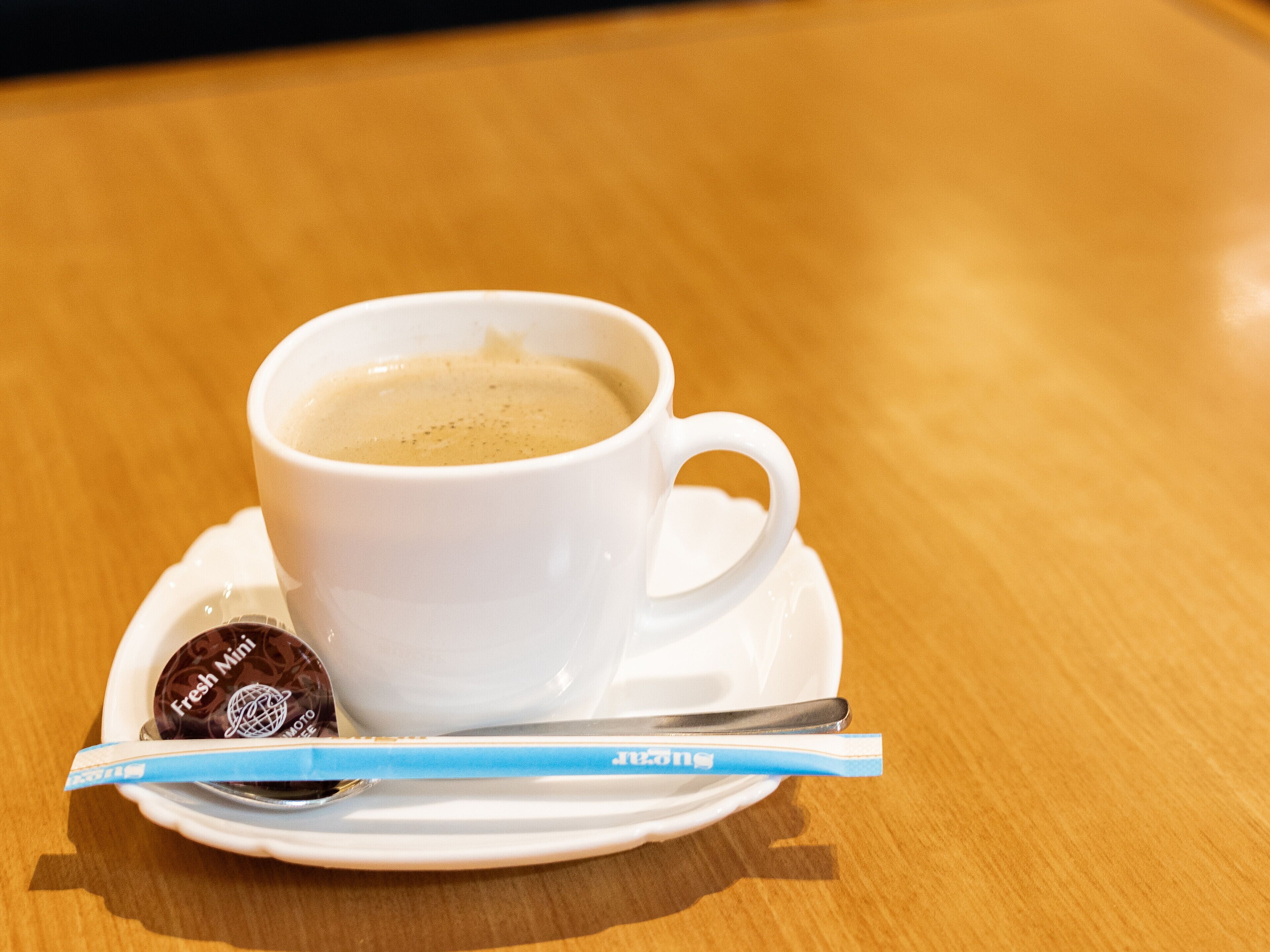 A white cup of coffee on a saucer with a spoon and a packet of Fresh Mini coffee.