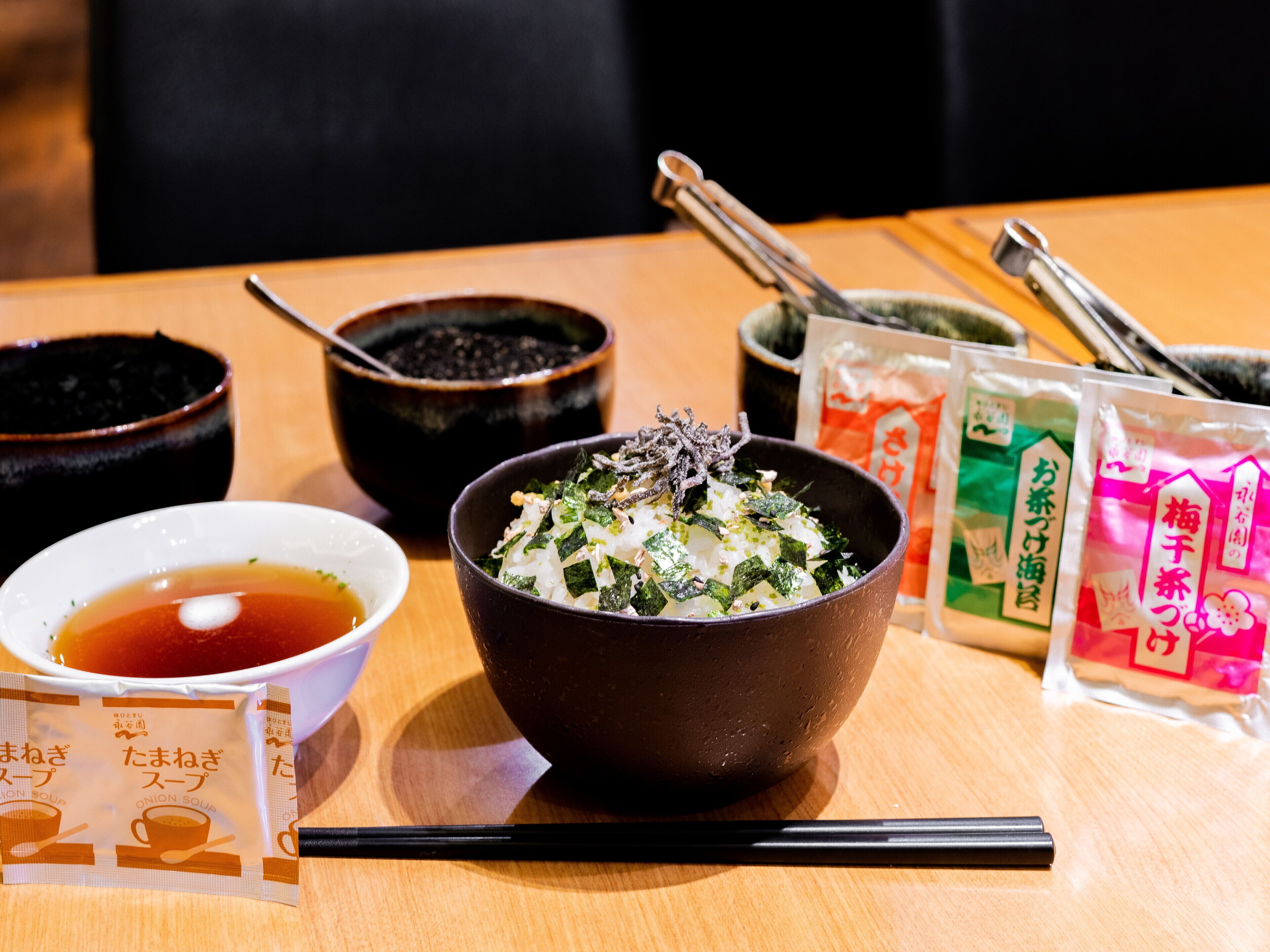 A table with a bowl of rice topped with seaweed and sesame seeds, a cup of tea, and packets of instant soup.