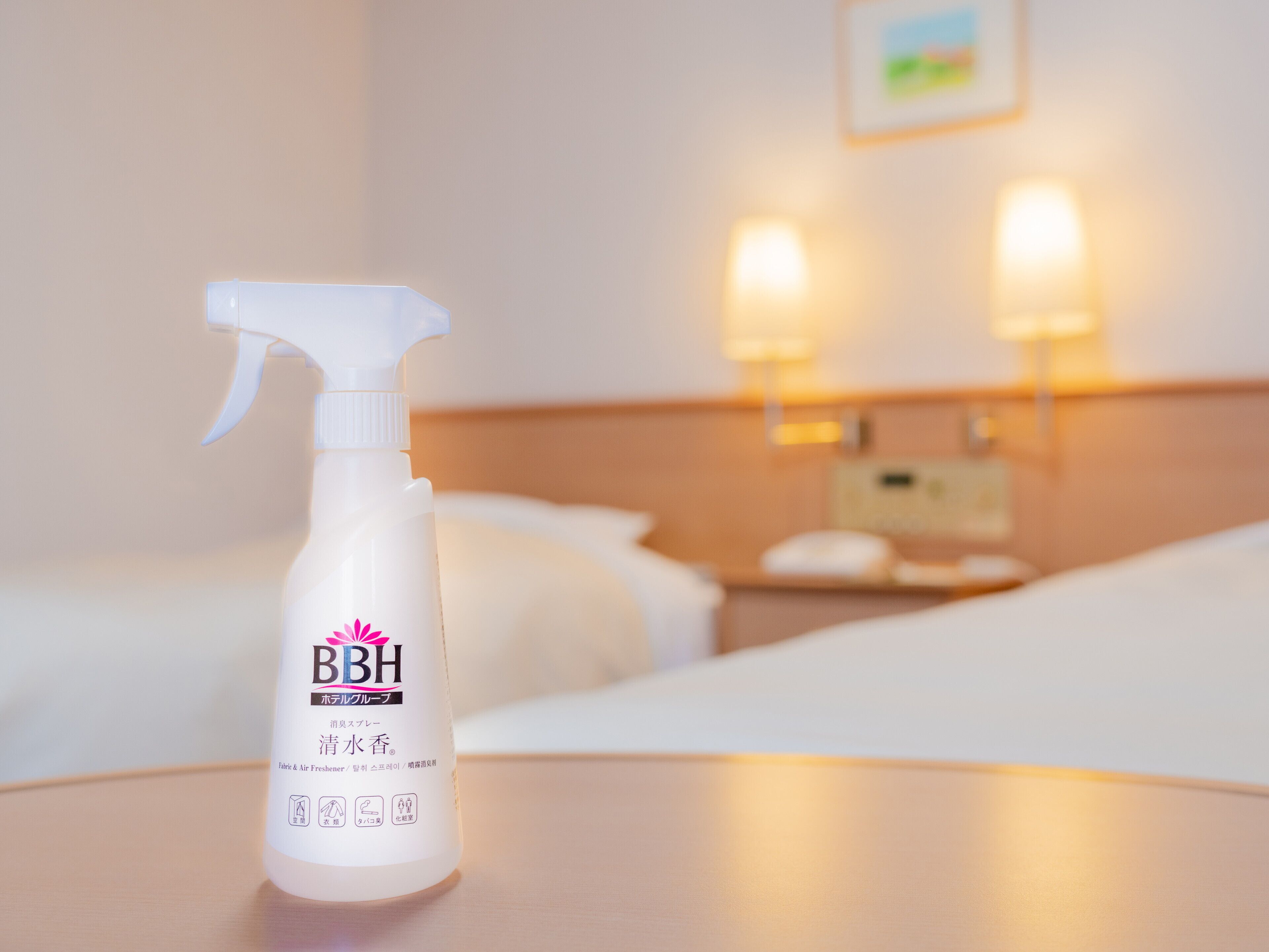 A hotel room with a bed, a nightstand, and a bottle of BBH fabric & air freshener on a table.