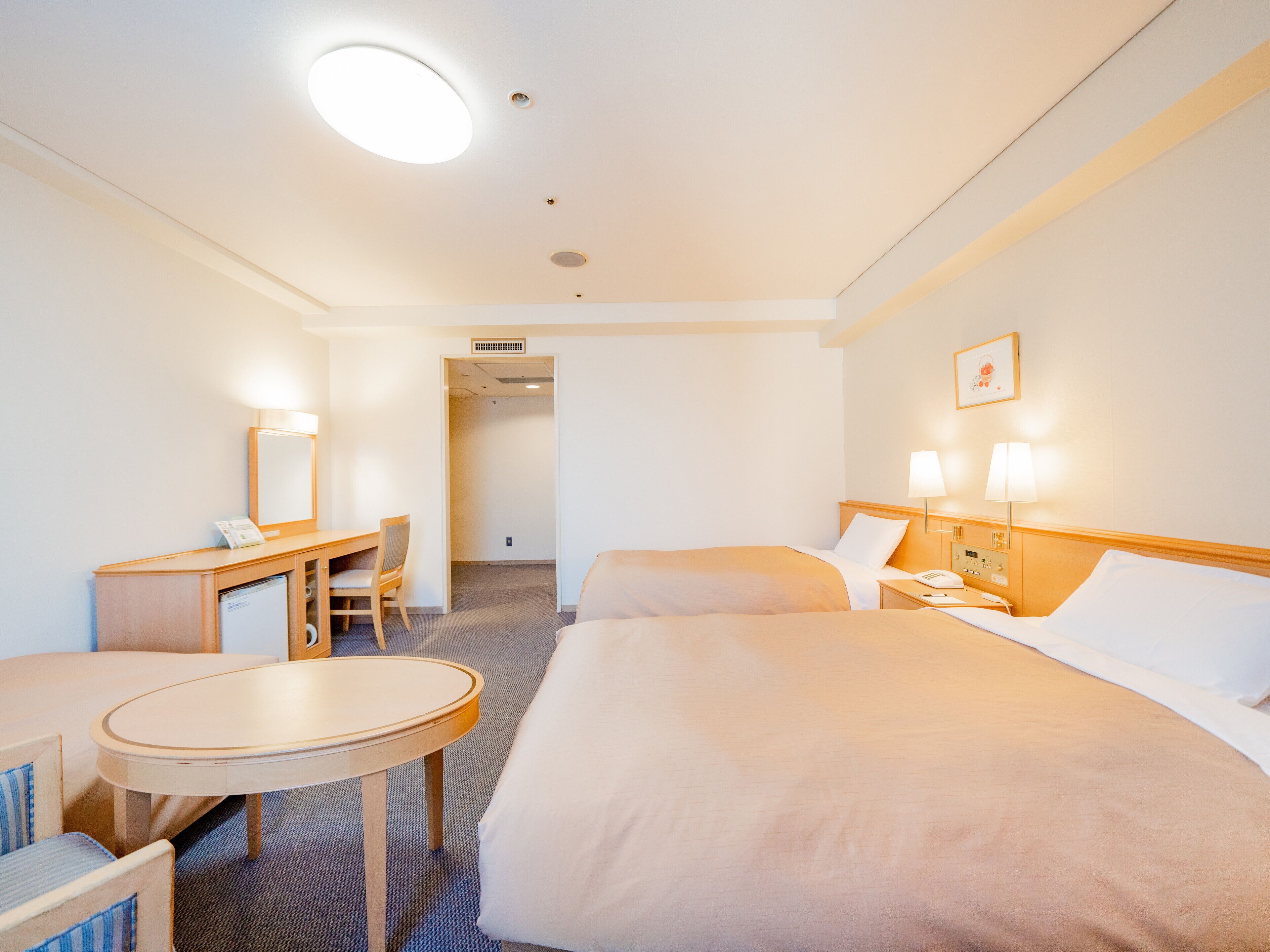 A hotel room with two beds, a desk, a chair, a small table, and a bedside lamp.