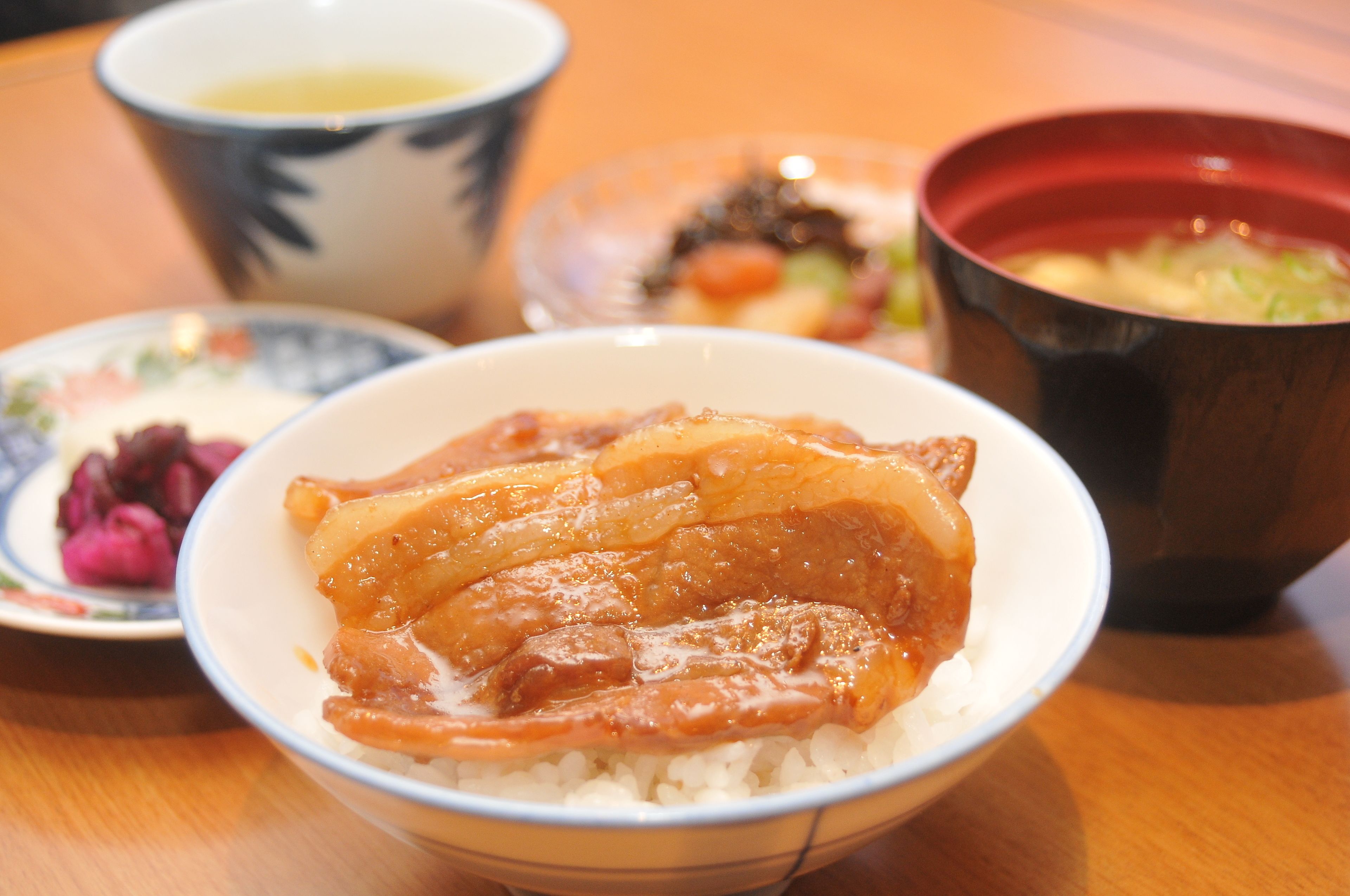 A bowl of rice topped with a glazed meat dish, accompanied by a small plate of pickled vegetables and a bowl of soup.