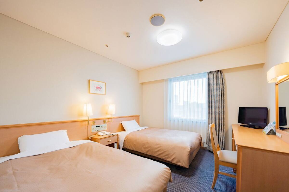A hotel room with two beds, a desk with a TV, a chair, and a window with curtains.