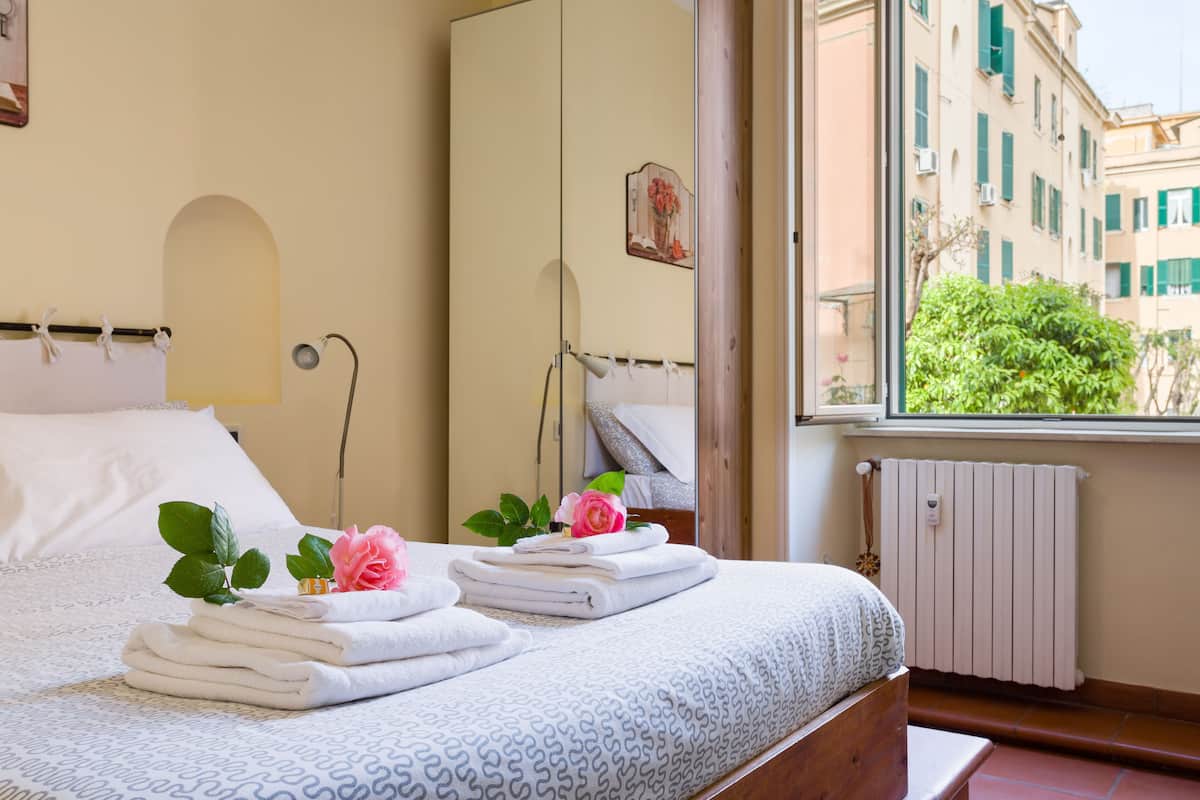 A neatly made bed with towels and a rose, a bedside lamp, and a window with a view of buildings.