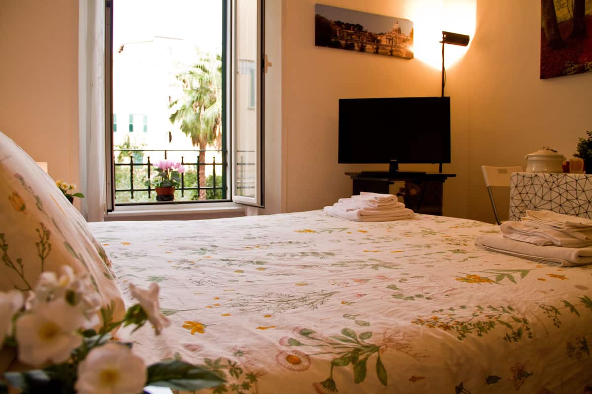 A bedroom with a bed, a television, a window with a view of palm trees, and a painting on the wall.