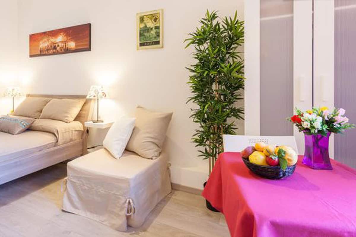 A hotel room with two beds, a nightstand with a lamp, a potted plant, and a table with a fruit basket.