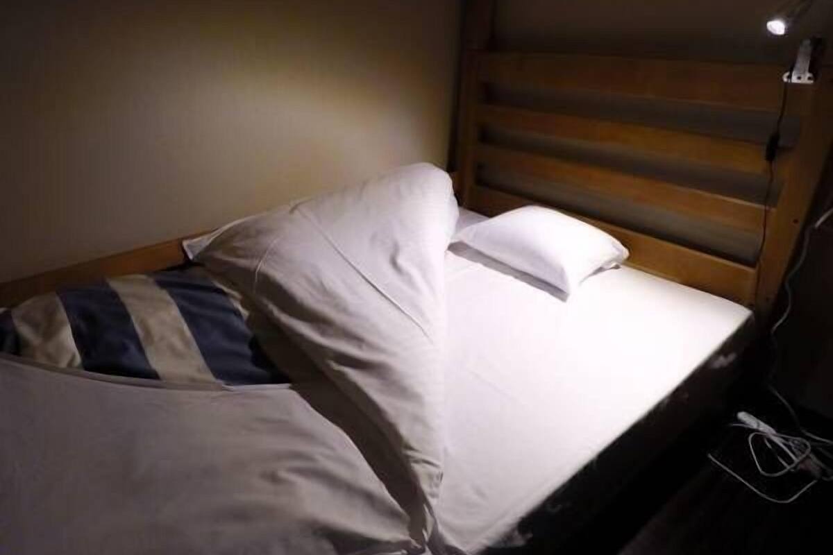 Um quarto pequeno e com pouca luz, com uma cama simples, cabeceira de madeira e uma tomada na parede.