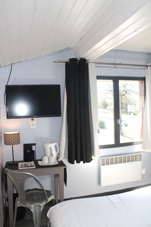 A hotel room with a bed, a desk with a computer, a flat-screen TV, a window with curtains, and a radiator.
