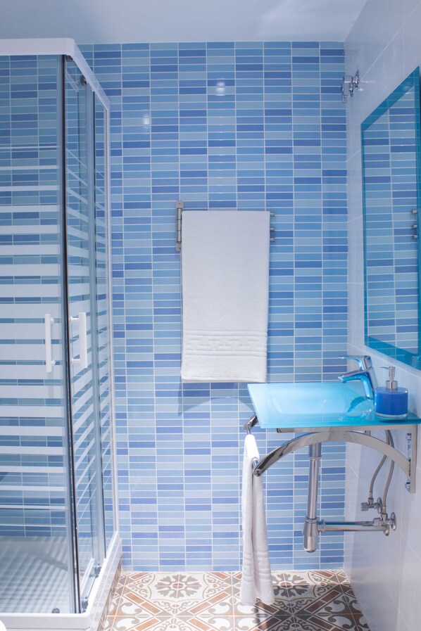 A bathroom with blue-tiled walls, a glass shower enclosure, a white towel hanging on the wall, and a blue sink with a mirror above it.