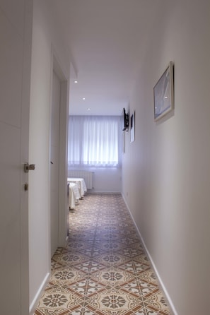 Un pasillo estrecho con baldosas de diseño, una cama con ropa blanca y una ventana con cortinas traslúcidas.