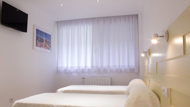 Habitación de hotel con dos camas, un televisor montado, una ventana con cortinas translúcidas e iluminación en la pared.