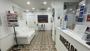 Una zona de recepción con suelo de baldosas, un sofá blanco, un mostrador de recepción con la etiqueta "BARCELONA" y un panel informativo en la pared.
