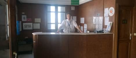 Una reception in un atrio d'albergo, con finiture in legno, una insegna per la reception e diverse locandine informative alle pareti.