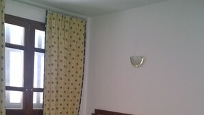 A room with a bed, a wooden chair, a table, a window with curtains, and a wall-mounted light fixture. - Hôtel L´Excellence (Ariana)