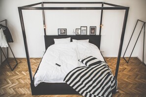 Uma cama com estrutura metálica preta, roupa de cama branca e piso de madeira em padrão herringbone.