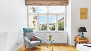 A room with a large window, a grey armchair, a small table with a plant, a lamp, and a bed with a yellow frame. - MGZN Basilica Rooms (Budapest)