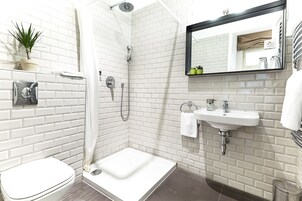 A modern bathroom with white brick walls, a white toilet, a shower with a handheld showerhead, a sink with a mirror above it, and a small potted plant on the wall.