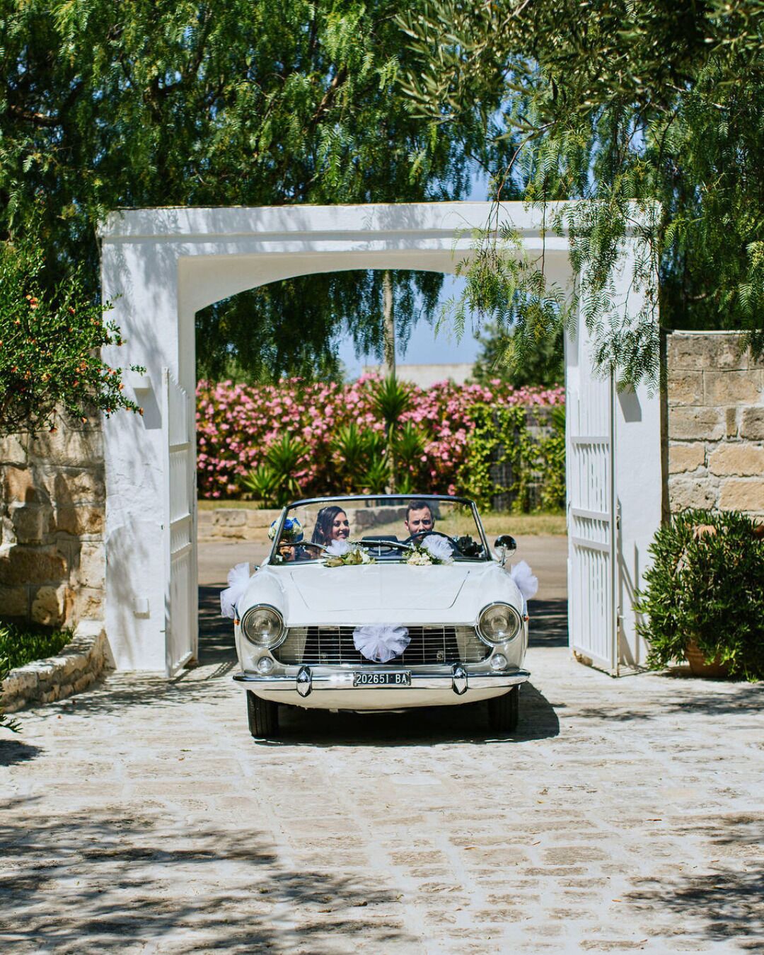 A white convertible car parked under a white archway with a stone wall and greenery in the background.
