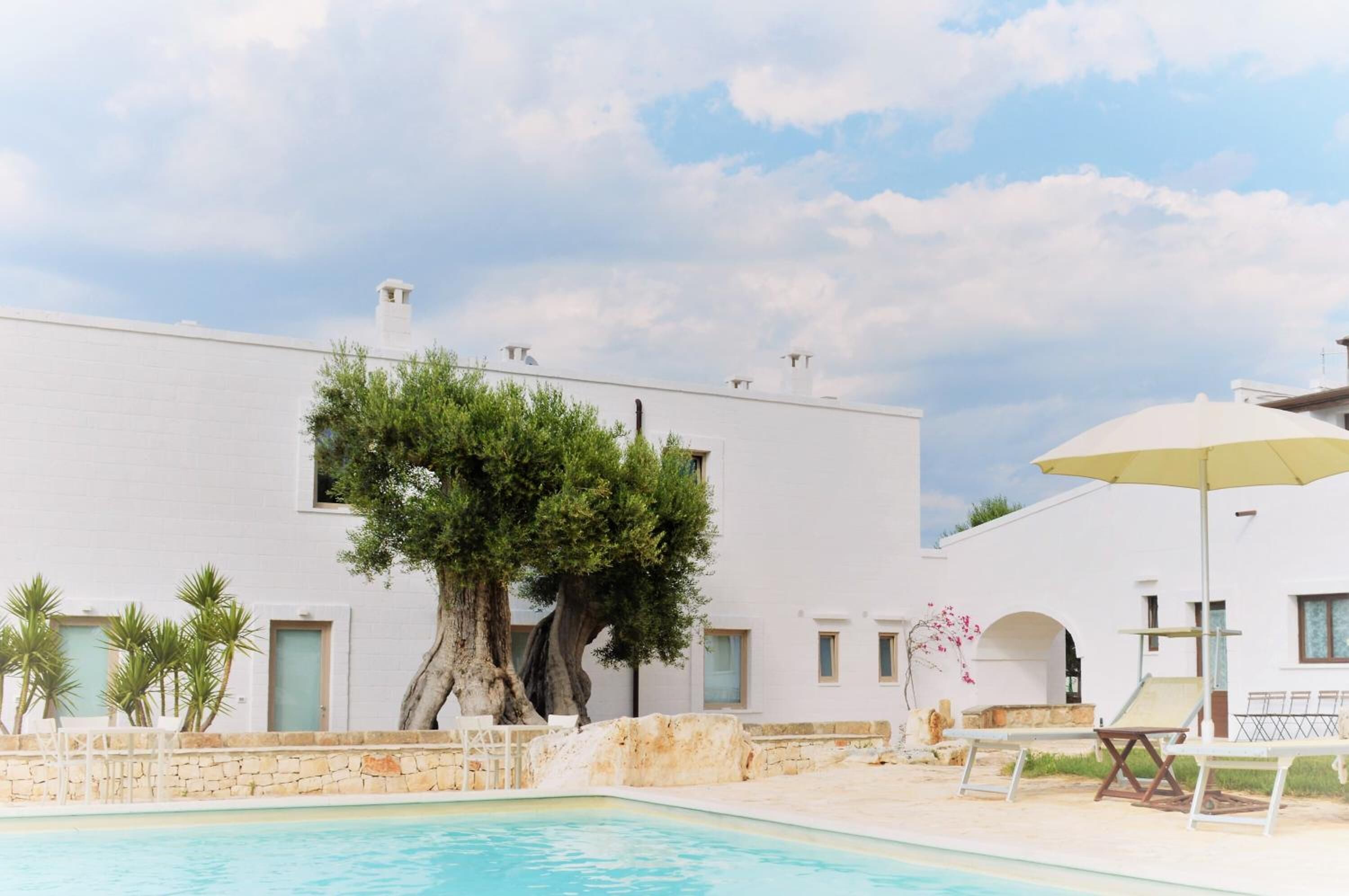 A pool area with a white building, a large tree, and a lounge chair with an umbrella.
