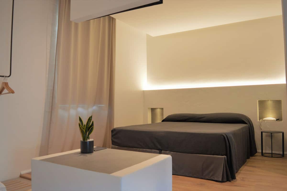 A modern bedroom with a bed, bedside tables, a potted plant, and a wall-mounted light fixture.