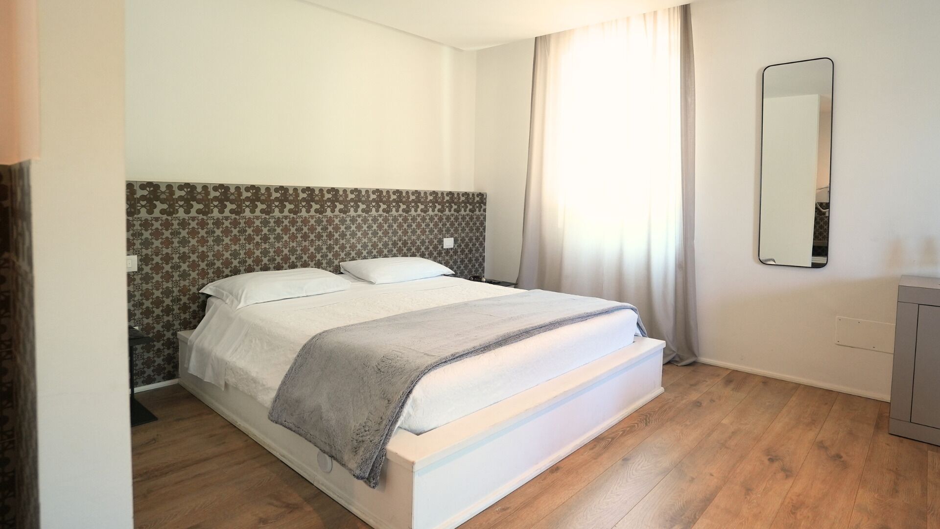 A bedroom with a bed, a headboard with a patterned design, a mirror, and a wooden floor.