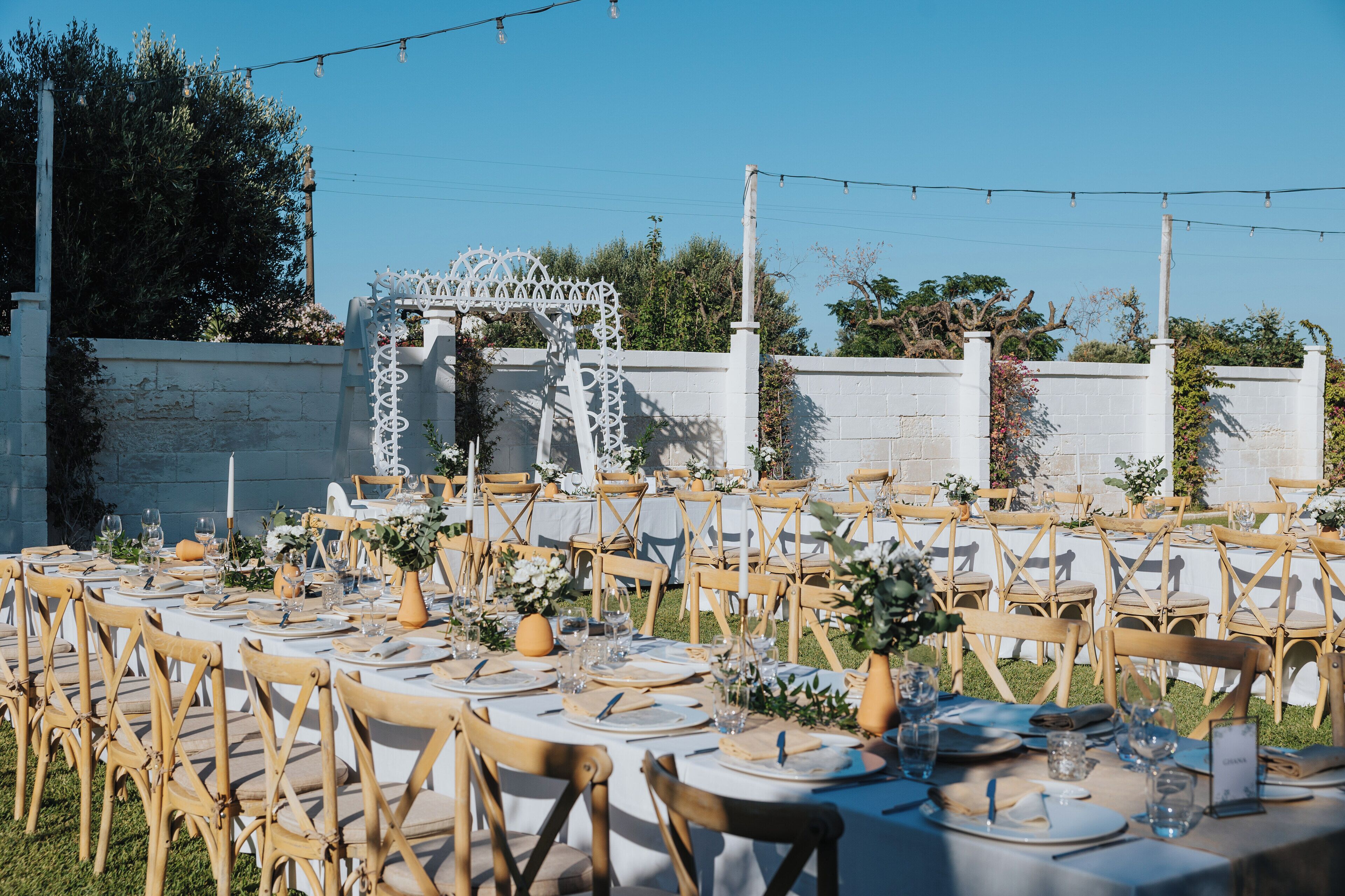 An outdoor dining area with white tables and wooden chairs, set against a white wall with a decorative archway.