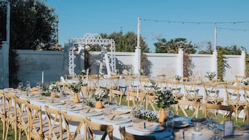 An outdoor dining area with white tables and wooden chairs, set against a white wall with a decorative archway.
