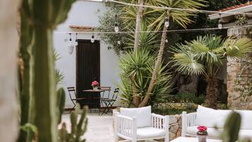 Un patio con muebles blancos, una mesa con un florero y diversas plantas, entre ellas cactus y palmeras.