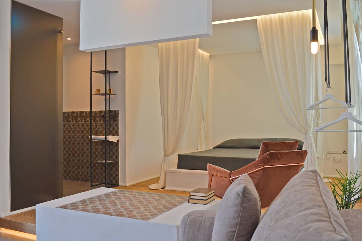 A modern hotel room with a bed, a seating area with a table, a bookshelf, and a bathroom with a shower and toilet.