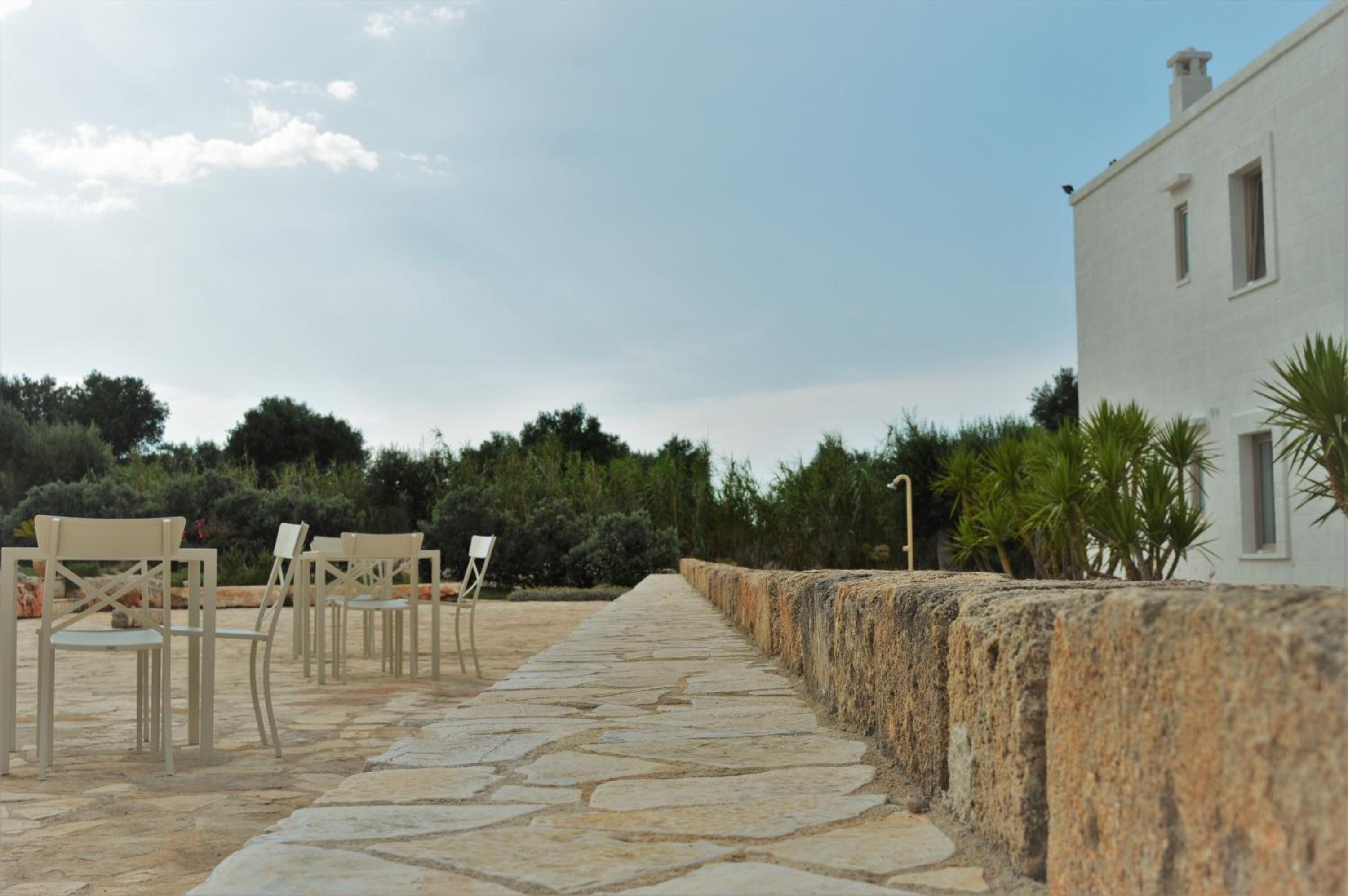 A stone pathway leading to outdoor seating with chairs and a table, surrounded by greenery and a building.