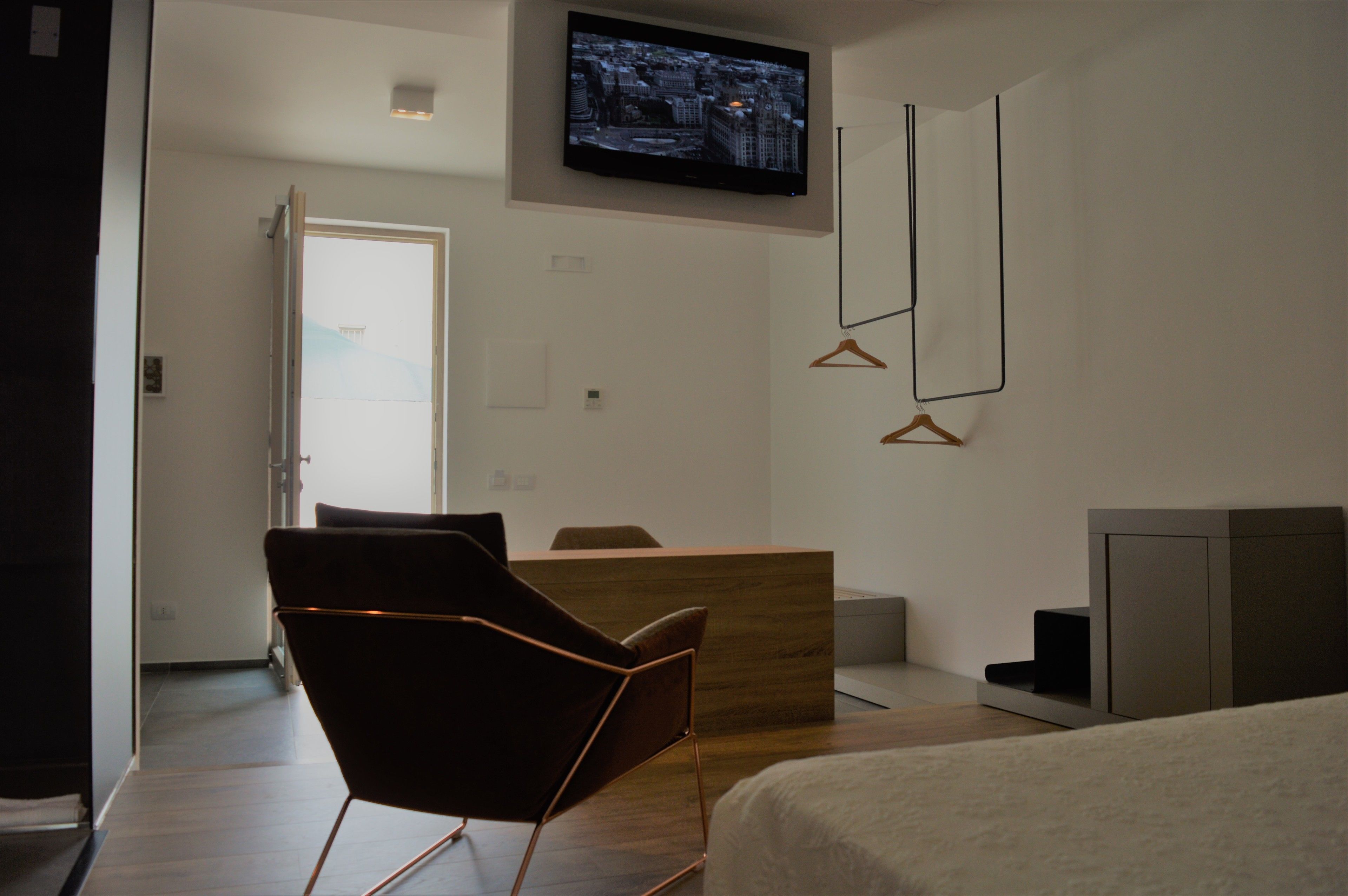 A modern hotel room with a wooden reception desk, a flat-screen TV mounted on the wall, a brown chair, and a bed with a white comforter.
