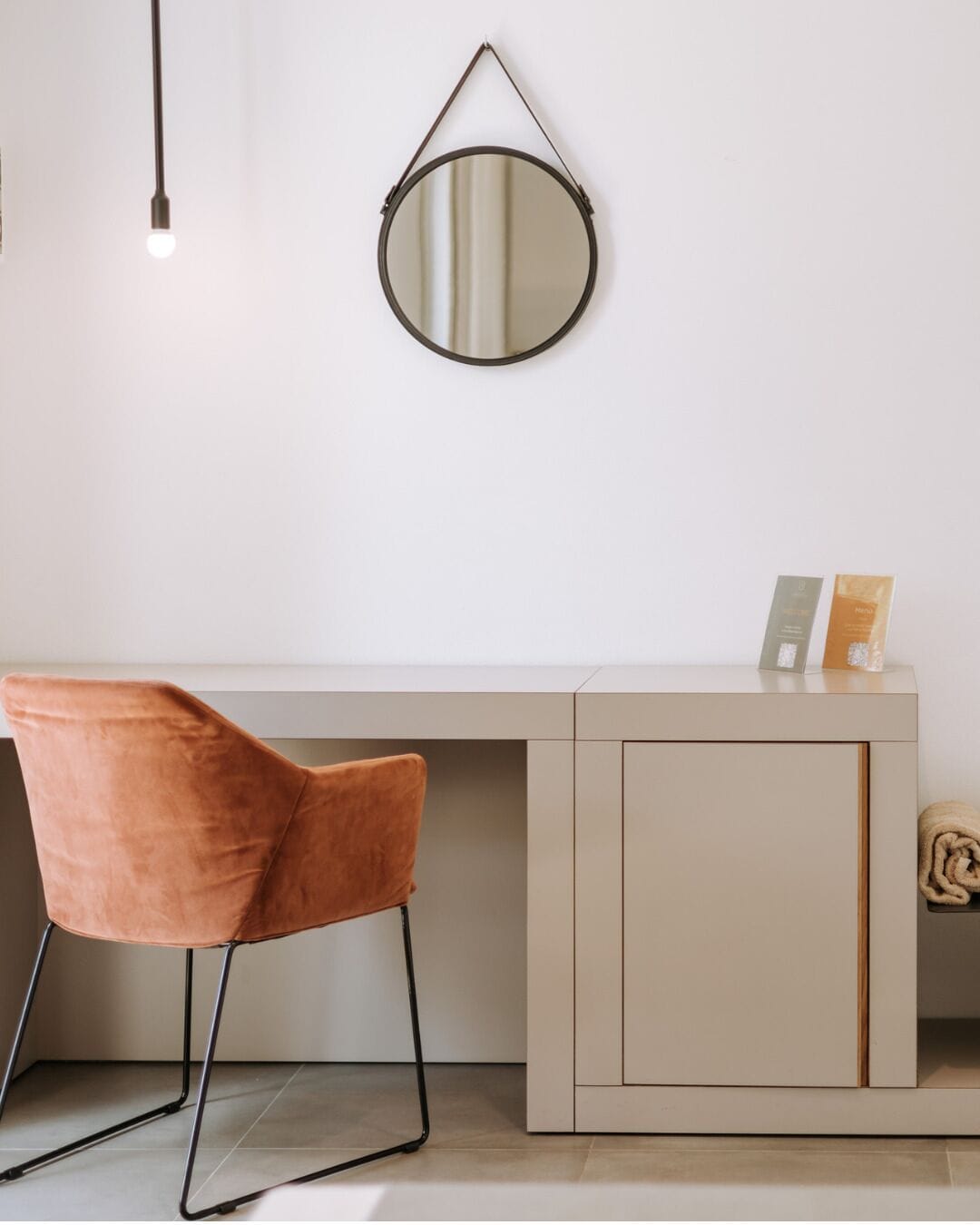Una habitación moderna con un escritorio blanco, un espejo redondo, una silla marrón y un mueble pequeño.