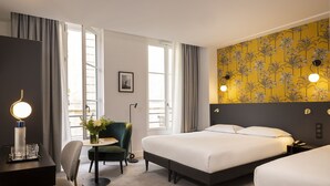 Una habitación de hotel con una cama, un escritorio con una silla, una mesita con un jarrón de flores y un papel tapiz floral amarillo en la pared.